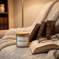 Candle labeled 'booklovers' on a couch with books and glasses, cozy atmosphere.